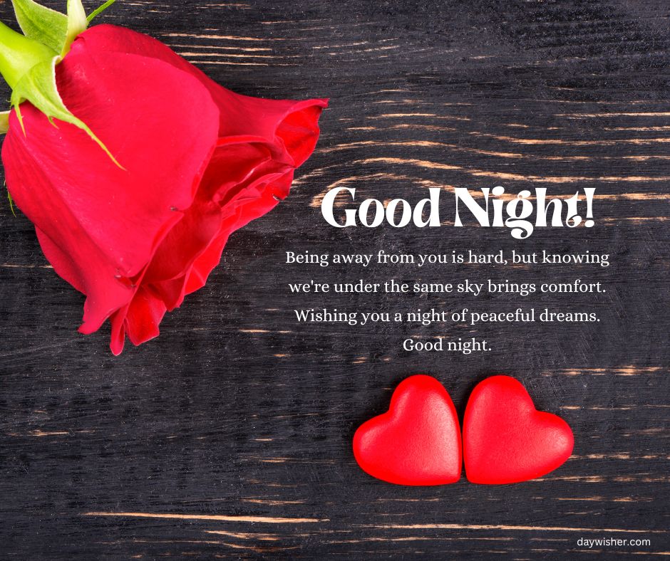 A red rose and two heart-shaped candies on a dark wooden surface with a text overlay that reads "Good night! Being away from you is hard, but knowing we're under the same sky brings comfort