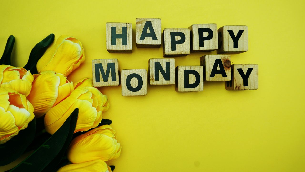 Wooden blocks spelling "happy Monday blessings" arranged next to a bunch of yellow tulips on a bright yellow background.