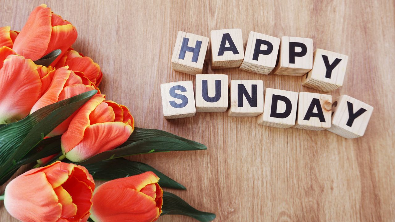 A cheerful arrangement of orange tulips beside wooden blocks spelling out "Happy Sunday Blessings" on a light wood surface.