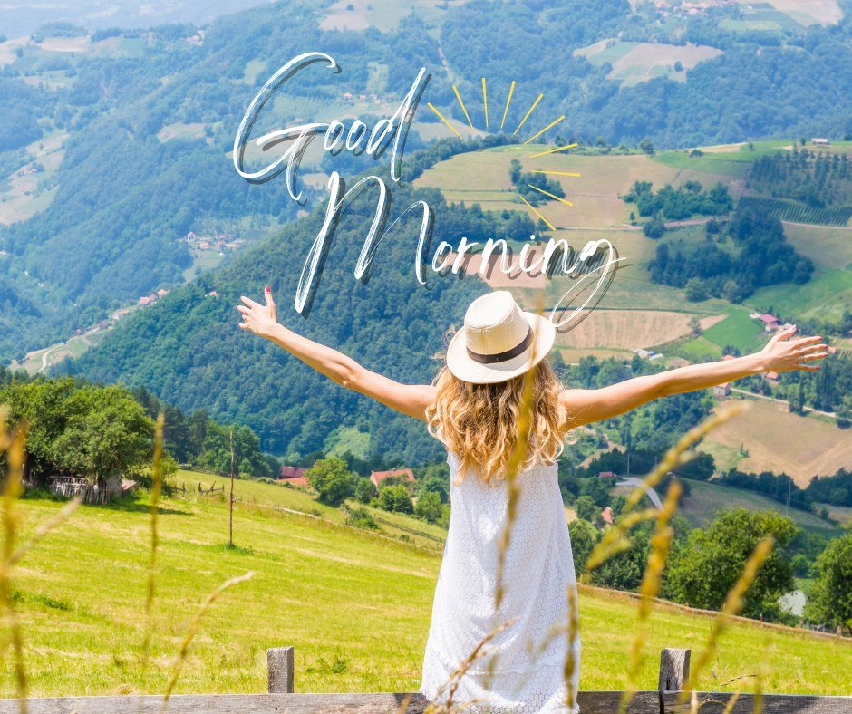 A vibrant good morning image featuring a woman in a white dress and sun hat, standing with her arms outstretched, overlooking a picturesque valley with lush green hills and fields. The text "Good Morning" is elegantly written in white with a sun illustration. The image captures the beauty and tranquility of the countryside, perfect for a refreshing morning greeting.