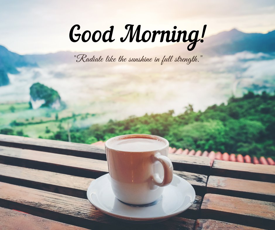 A cup of coffee on a wooden table overlooking a scenic landscape with misty mountains and lush greenery. The text "Good Morning!" is displayed above the quote "Radiate like the sunshine in full strength." This image beautifully captures the peacefulness of a morning with natural sunshine, perfect for sharing positive and inspiring vibes.