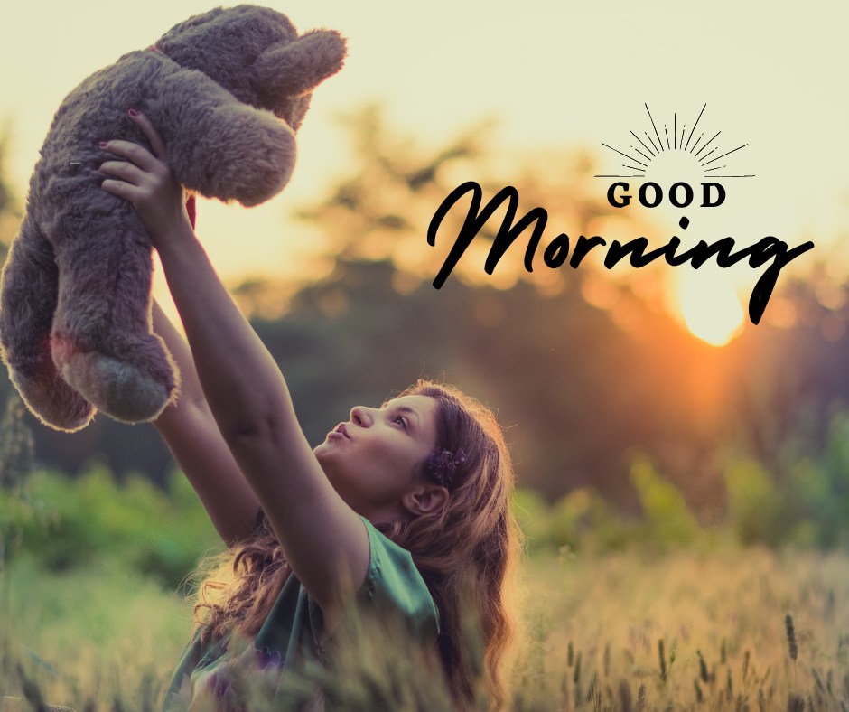 A woman holding up a good morning teddy bear in a field at sunrise. The image features the text "Good Morning" with the sun rising in the background, creating a warm and uplifting scene.