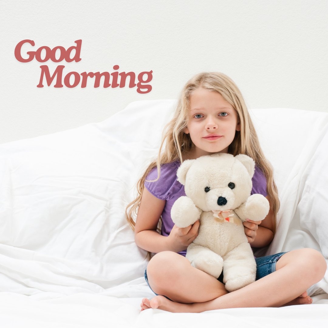 A young girl sitting on a bed, holding a white good morning teddy bear. The image features the text "Good Morning" in a warm, welcoming font, creating a cozy and sweet morning scene.