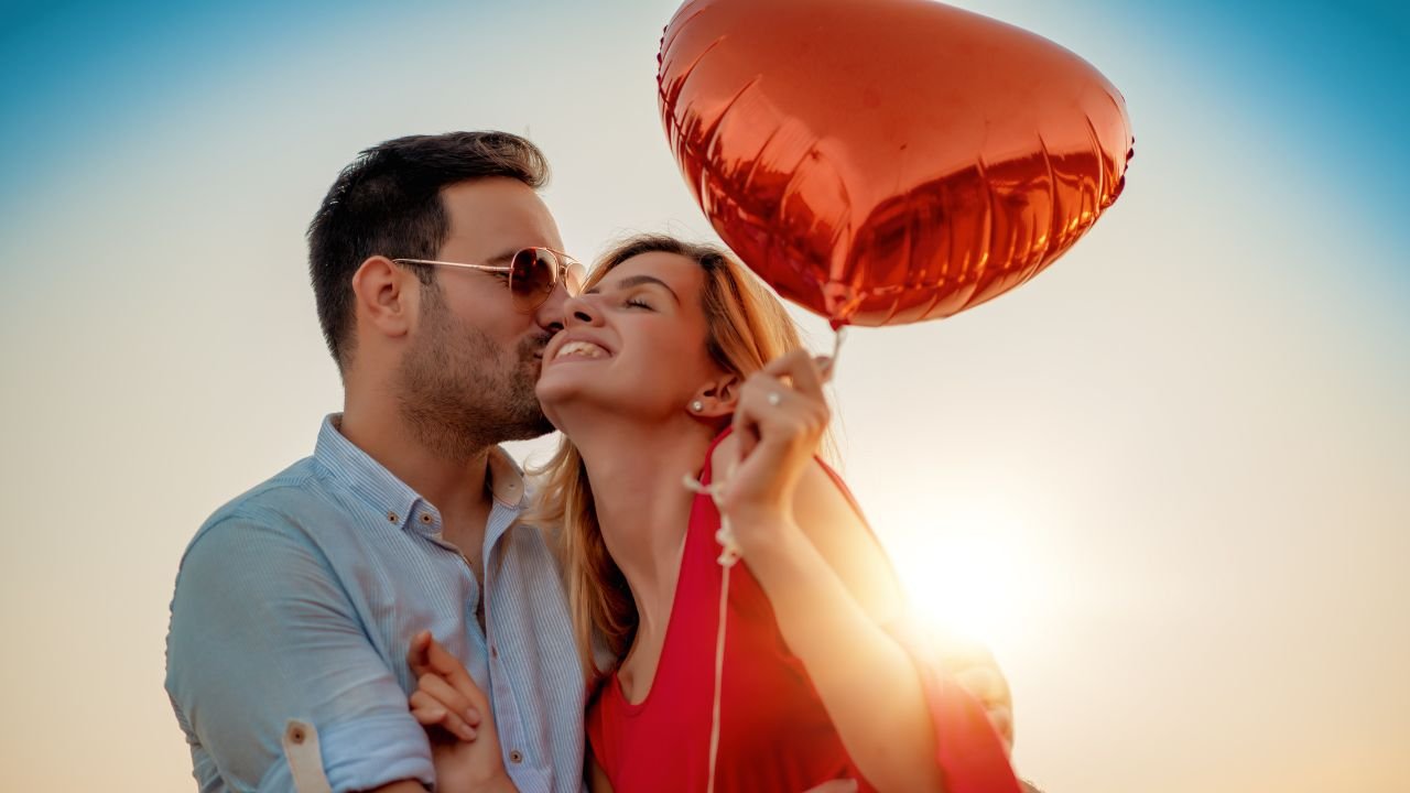 A couple embraces while holding a heart-shaped balloon, smiling during a sunset, showcasing affection and celebrating their Happy Monthsary.