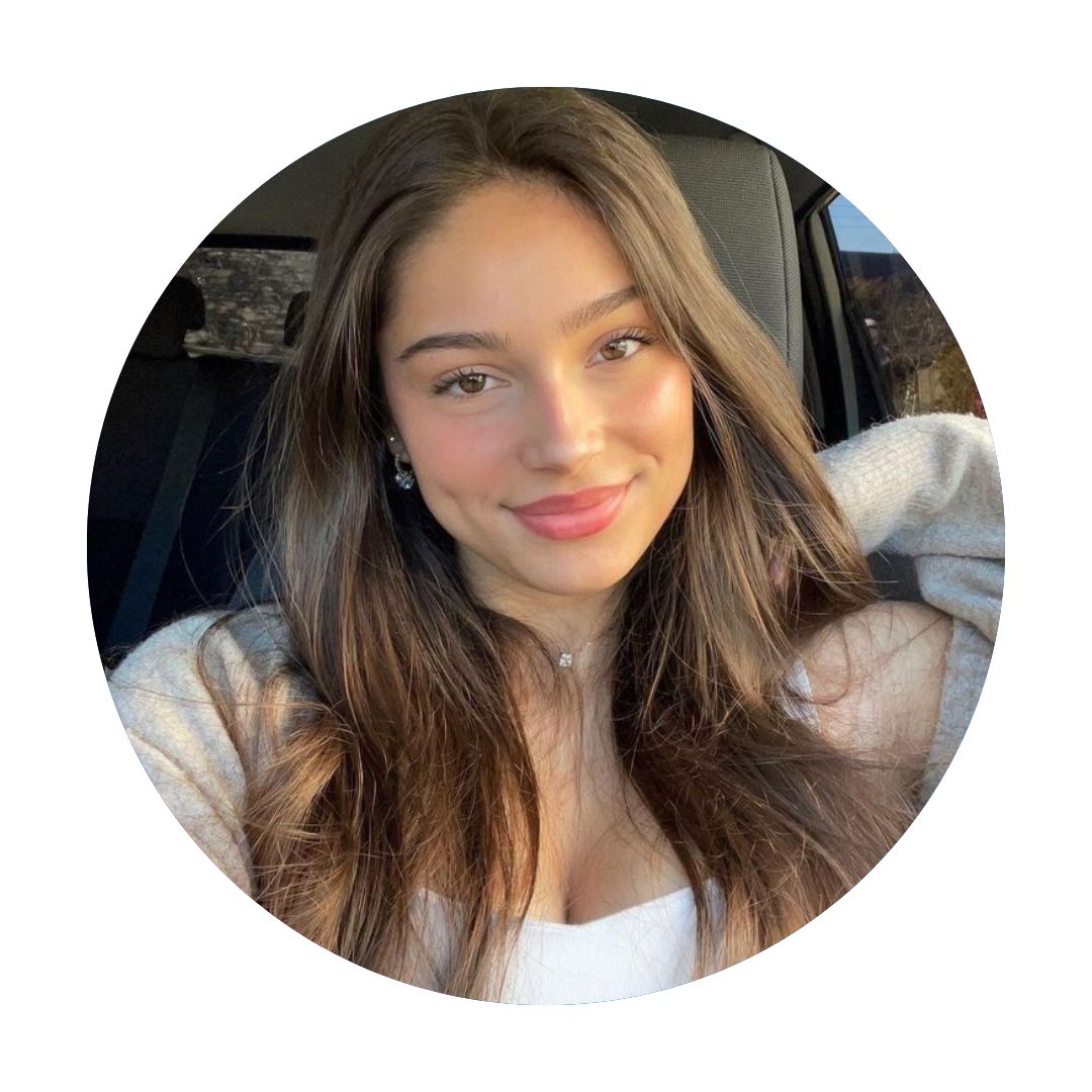 A close-up portrait of a smiling young woman with long brown hair sitting in a car, sunlight illuminating her face, dressed in a light sweater, with a seatbelt visible.